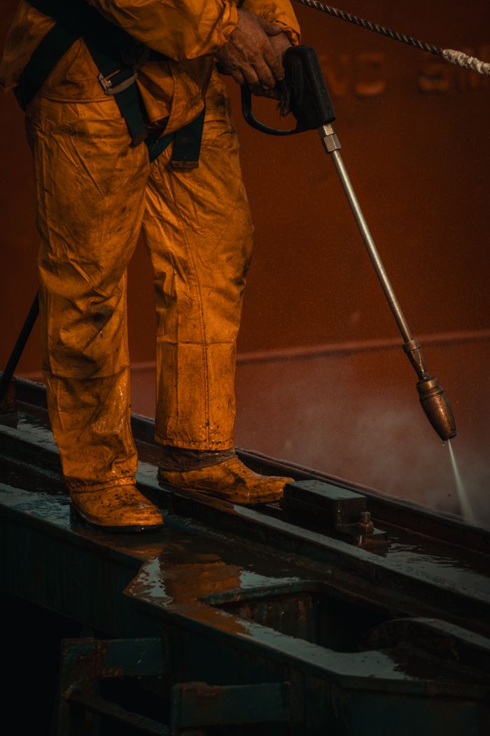 A worker in yellow safety gear is using a pressure washer on industrial equipment.